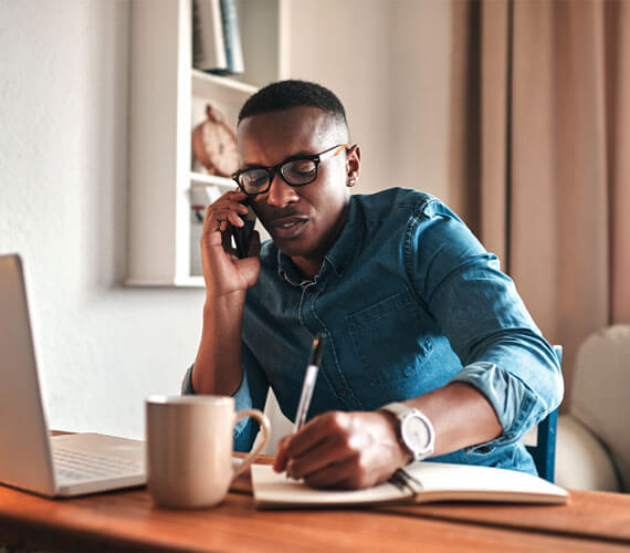 Man working at a wooden desk with a laptop, a mug, and a notebook while talking on a mobile phone