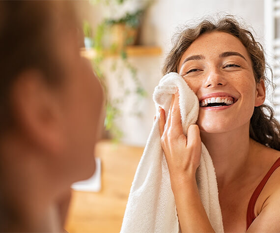Smiling woman gently drying her face with a soft white towel in front of a mirror.
