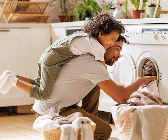 Father loading a washing machine while his young child rides piggyback on his back