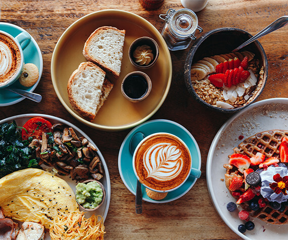 Breakfast table featuring sourdough toast, an egg and mushroom plate, waffles with fruit, and a latte