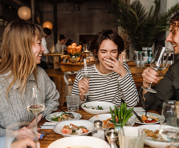 Group of friends laughing and drinking wine while having a meal together at a restaurant.